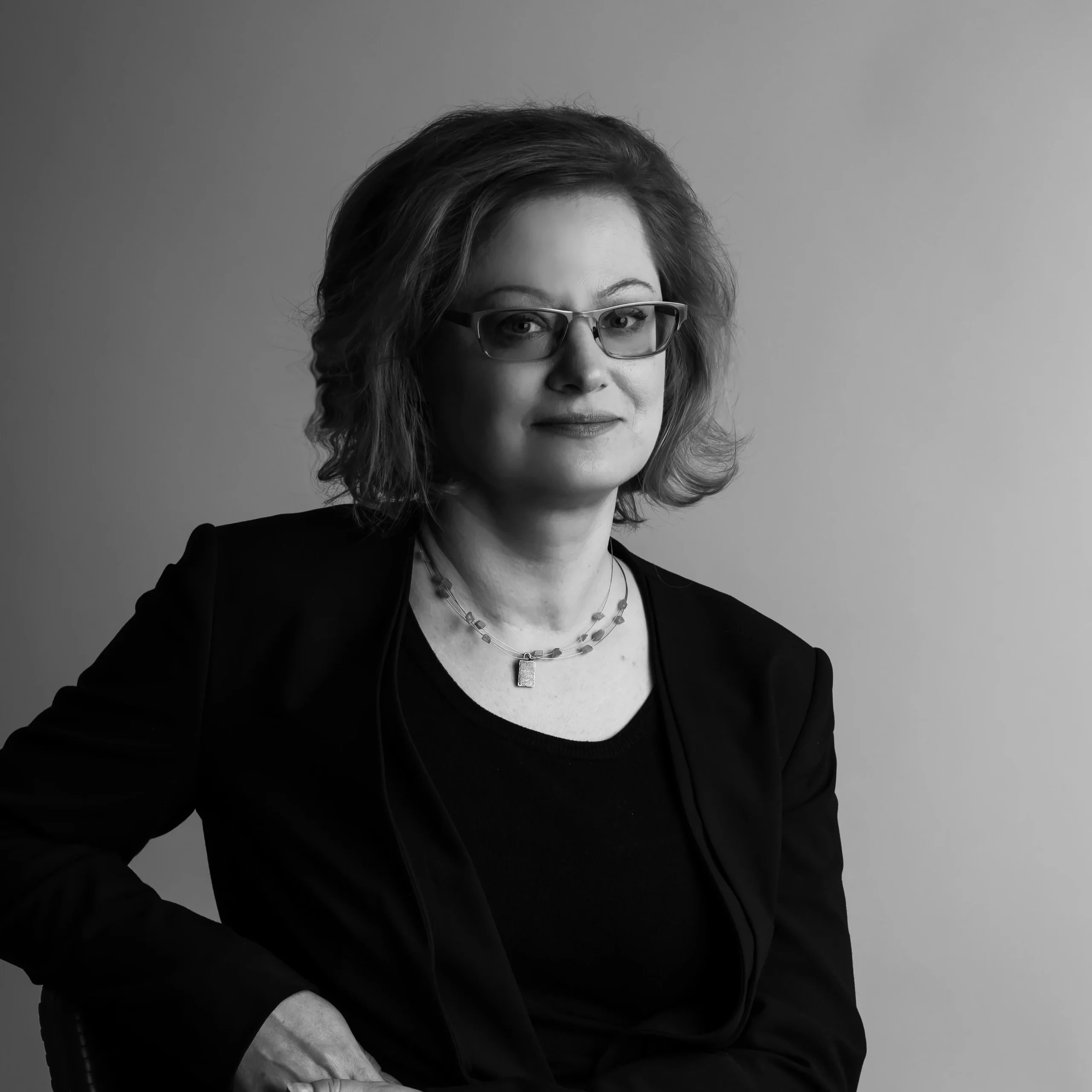 Black and white photo: a woman with glasses, short light hair, wearing a black blazer and top, sitting and posing with a gentle smile.