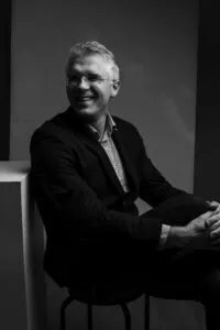 A happy professor with glasses and a suit is posing for a picture with his hands clasped around his one leg.