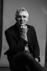 A professor with glasses in a suit is sitting on a chair, resting his chin on his hand.