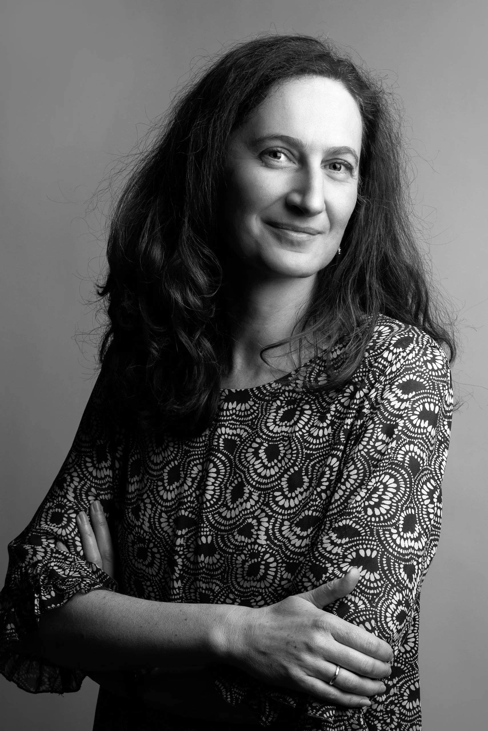 A happy dark, long-haired professor is posing for a camera wearing a printed shirt, with her hands clasped on her chest.