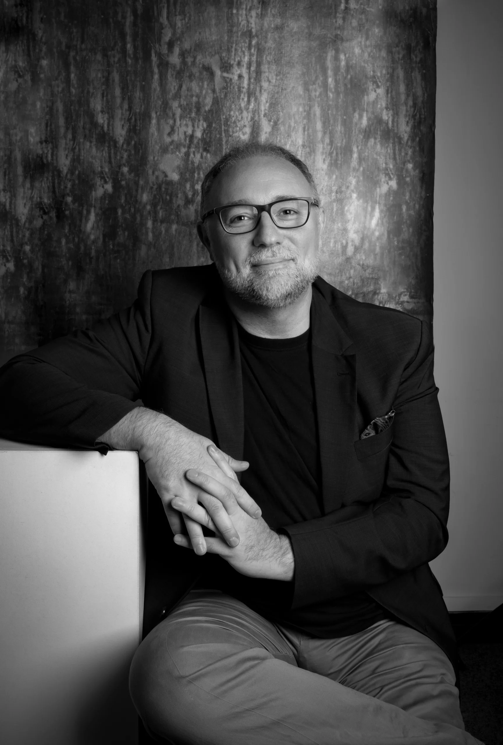 a professor in a suit is posing for a picture. He is leaning on a white box with his hands clasped.