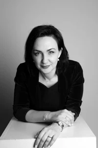 Dark-haired professor is posing for a camera, while leaning on a white box with her arms.