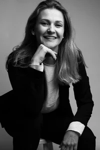 A professor with long light hair and in a suit is posing for a picture, resting her chin on her right hand.