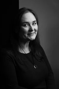 A happy dark-haired professor, wearing a dark shirt, is posing for a picture.