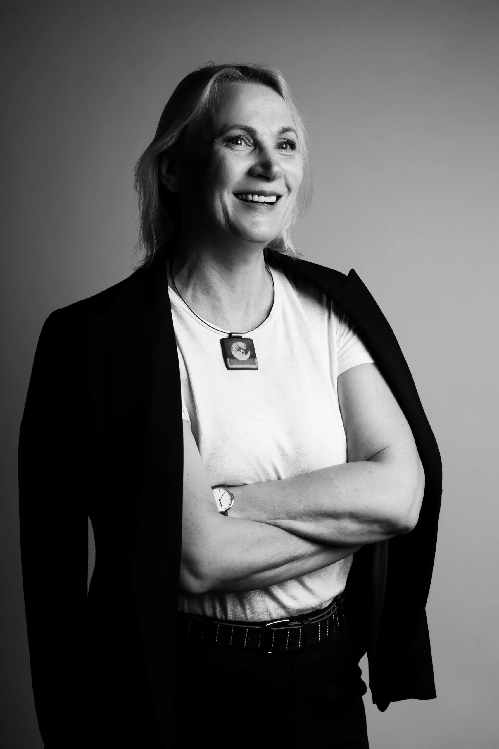 Happy, light-haired professor with a white t-shirt and a black blazer is posing for a picture with her arms crossed on her chest.