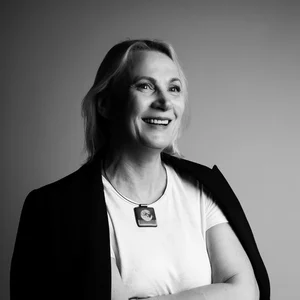 Happy, light-haired professor with a white t-shirt and a black blazer is posing for a picture with her arms crossed on her chest.