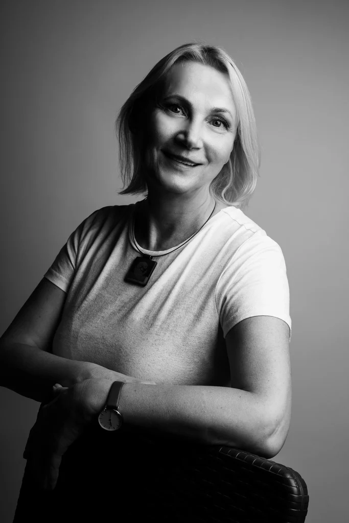 Happy, light-haired professor, with a white t-shirt, is posing for a picture while leaning on a chair with her arm.