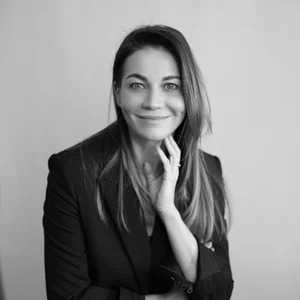 A black and white portrait photograph of a smiling woman with long hair, wearing a black blazer. She is looking directly at the camera, gently resting her face on her hand.
